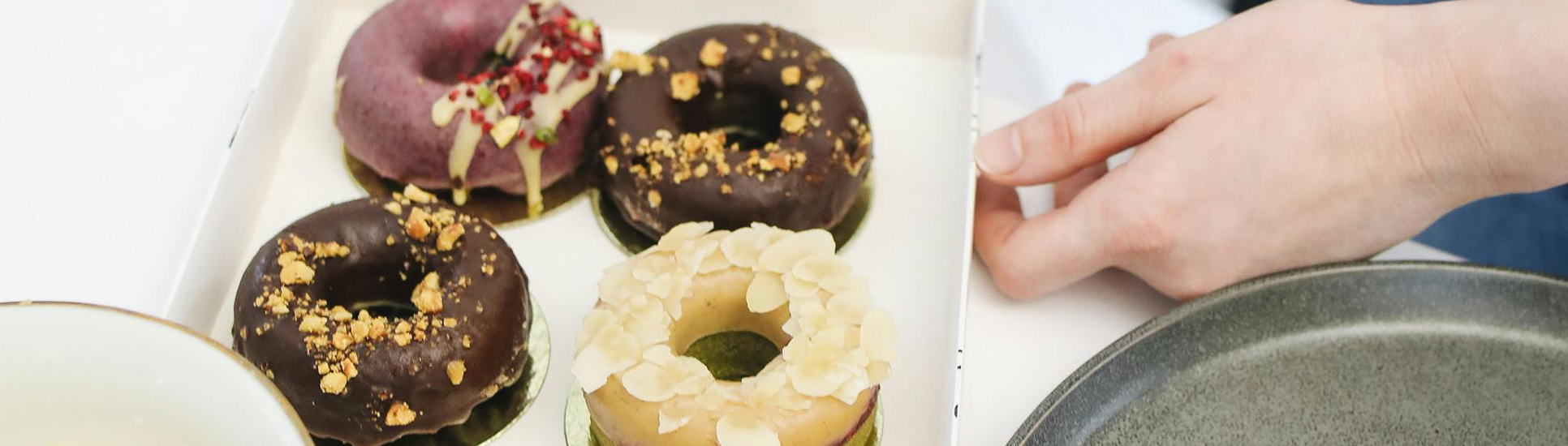 Boîte à emporter contenant quatre donuts artisanaux colorés, prête à être refermée par une main qui tient la boîte.