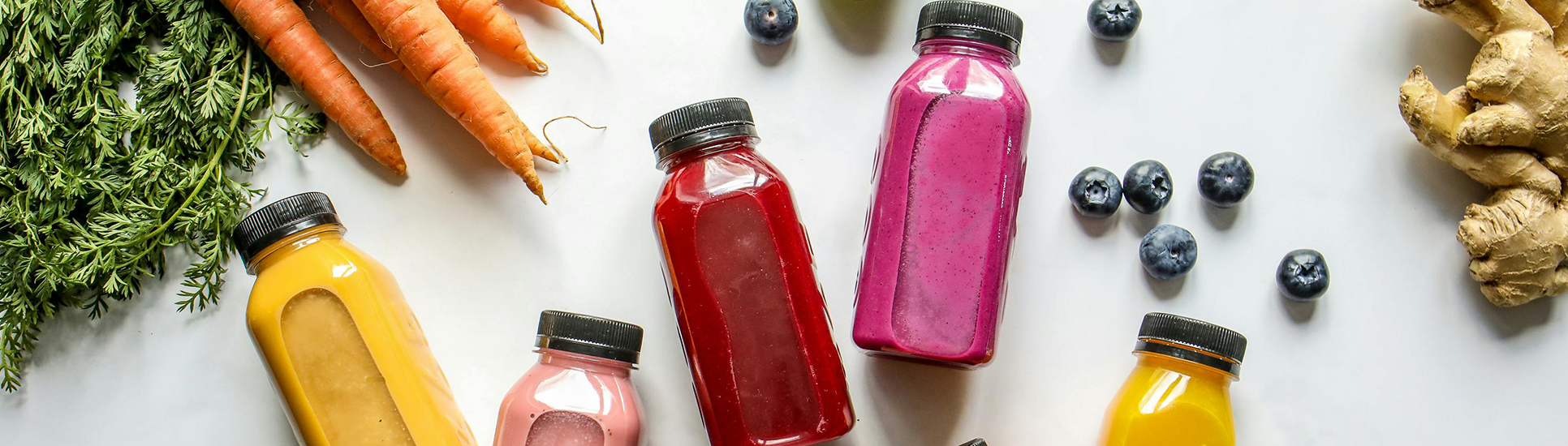 Bouteilles de smoothies colorés à base d'ingrédients frais comme carottes, gingembre, myrtilles, disposés sur une table blanche.