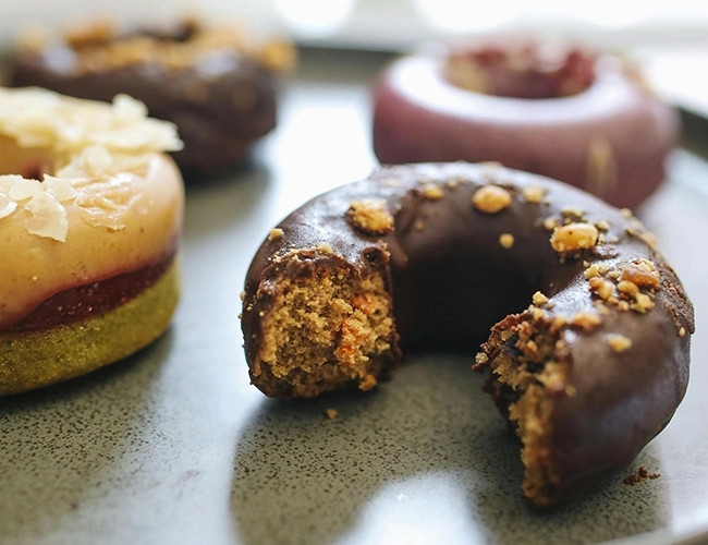 Donut au chocolat croqué, garni de noisettes, accompagné d'autres donuts artisanaux colorés en arrière-plan.